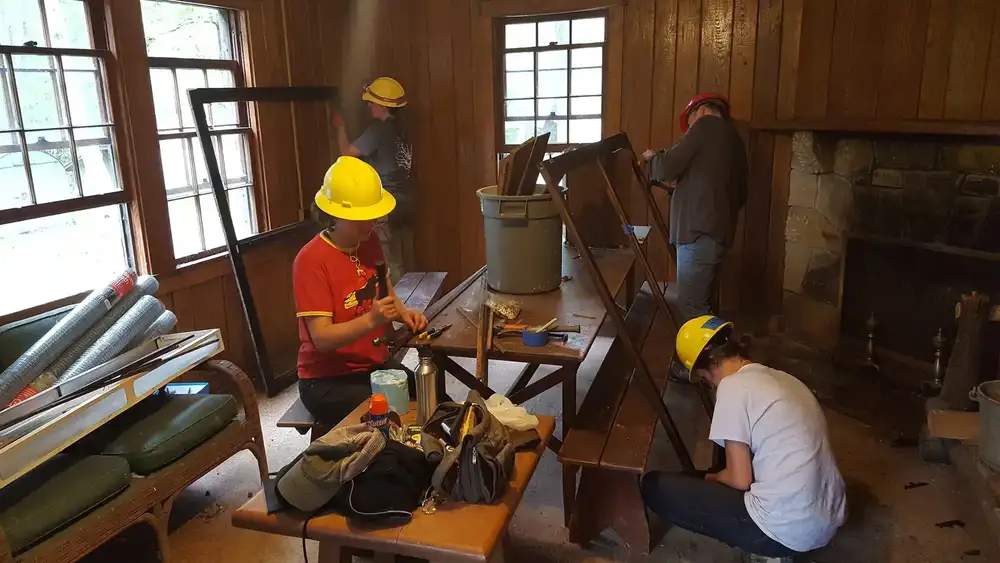 a crew in hard hats rebuilding wooden screen frames, with rolls of screen mesh on a bench
